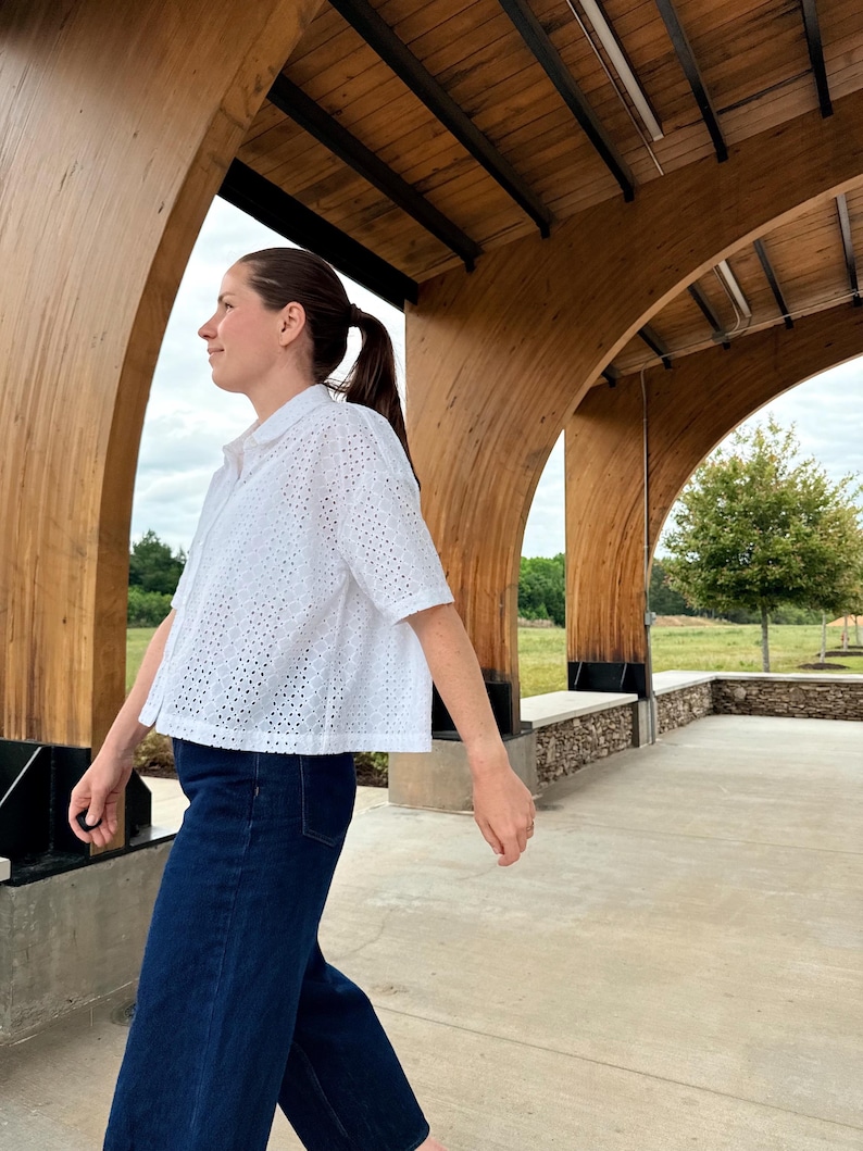 May include: A white eyelet shirt with short sleeves and a collared neckline, worn with dark blue denim trousers. The shirt features a perforated pattern. The person is walking under a wooden structure with a concrete floor.