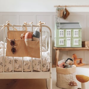 May include: A white metal crib with a yellow and white gingham fabric hanging organizer. The organizer has a star, sunglasses, and stuffed toys inside. The crib has a floral patterned sheet.