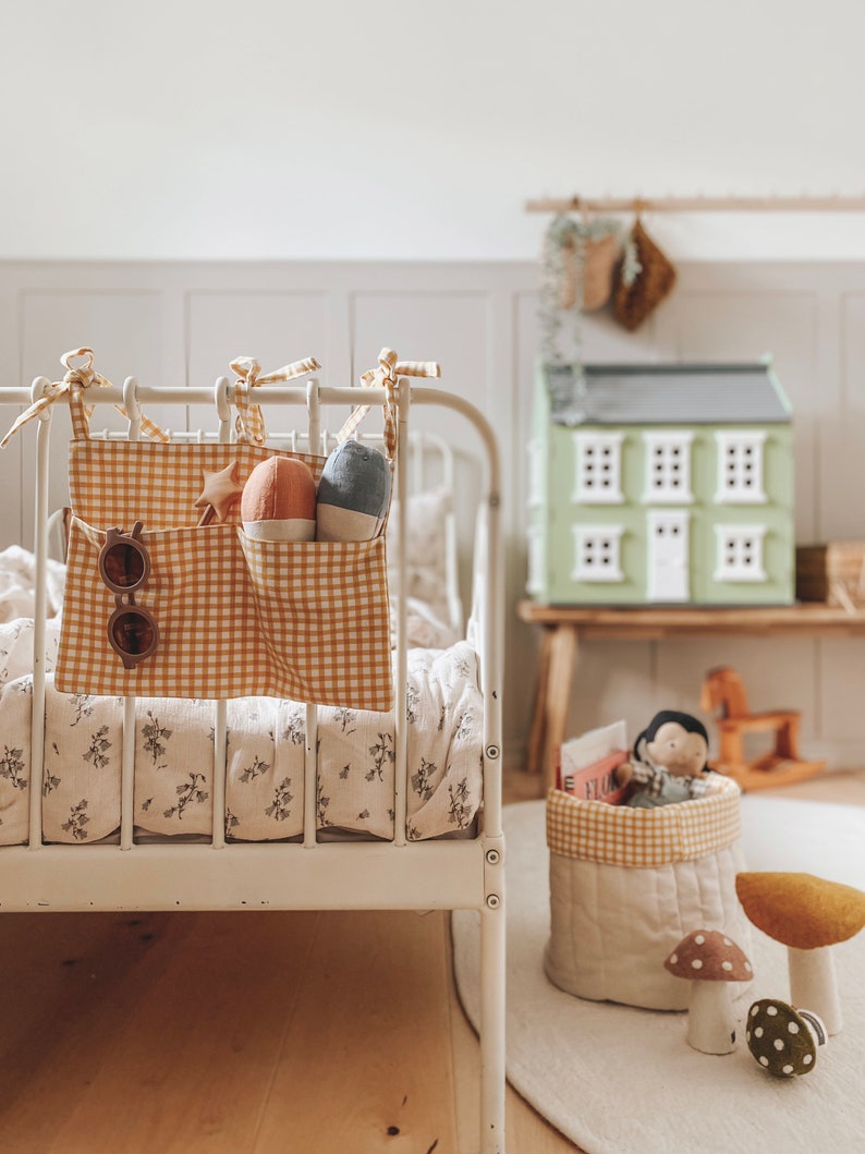 May include: A white metal crib with a yellow and white gingham fabric hanging organizer. The organizer has a star, sunglasses, and stuffed toys inside. The crib has a floral patterned sheet.
