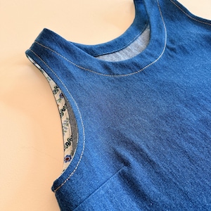 May include: Close-up of a sleeveless denim dress. The dress is a medium blue color with a round neckline and a contrasting floral print lining the armholes. The seams are highlighted with light-colored stitching.