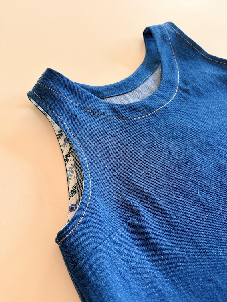 May include: Close-up of a sleeveless denim dress. The dress is a medium blue color with a round neckline and a contrasting floral print lining the armholes. The seams are highlighted with light-colored stitching.