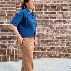 May include: A blue denim short-sleeved shirt with two front pockets and tan corduroy trousers. The outfit is completed with red shoes. The background is a brick wall.