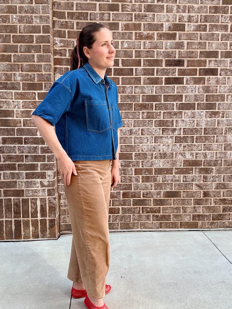 May include: A blue denim short-sleeved shirt with two front pockets and tan corduroy trousers. The outfit is completed with red shoes. The background is a brick wall.