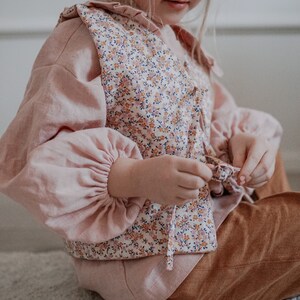 Pu&ograve; includere: Una bambina che indossa una blusa rosa con un gilet a stampa floreale. Il gilet ha una chiusura a cravatta e la bambina sta regolando la cravatta. Indossa pantaloni marroni.