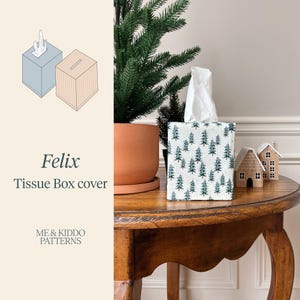 May include: A rectangular tissue box cover featuring a repeating pattern of green pine trees on a white background. The cover is placed on a wooden surface next to a small decorative house and a potted evergreen plant. The text Felix Tissue Box cover is visible.