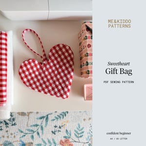 May include: A red and white gingham heart-shaped gift bag with a loop handle. The image also shows a sewing machine, a floral patterned box, and the text "Sweetheart Gift Bag PDF Sewing Pattern" and "ME&KIDDO PATTERNS".