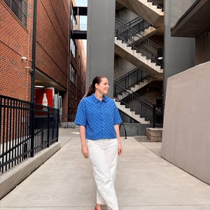 May include: A person wearing a blue patterned short-sleeved shirt, white trousers, and red flats stands on a concrete walkway. The shirt has a small geometric pattern. The background includes brick buildings and a metal staircase.