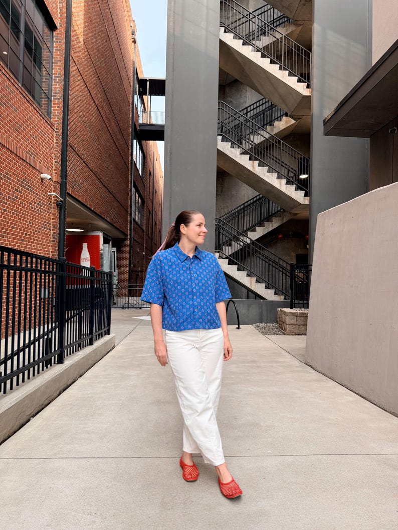 May include: A person wearing a blue patterned short-sleeved shirt, white trousers, and red flats stands on a concrete walkway. The shirt has a small geometric pattern. The background includes brick buildings and a metal staircase.