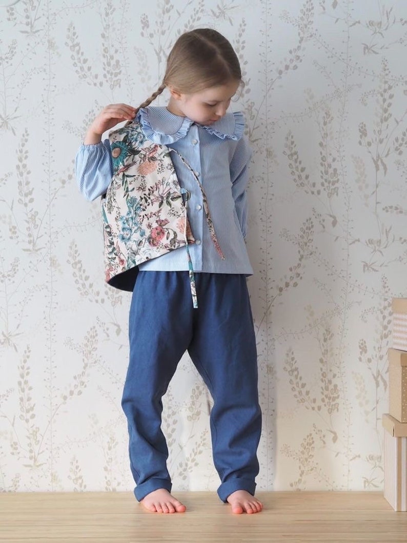 Pu&ograve; includere: Una giovane persona indossa una camicia abbottonata blu chiaro con colletto bianco e un gilet a stampa floreale. La camicia ha delle strisce bianche e il gilet ha un colletto arricciato. La persona indossa pantaloni blu scuro e ha i capelli intrecciati.
