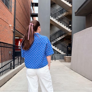 May include: A person wearing a blue patterned short-sleeved shirt and white trousers. The shirt features a floral design. The person's hair is styled with a pink ribbon. The background includes brick buildings and a concrete walkway.