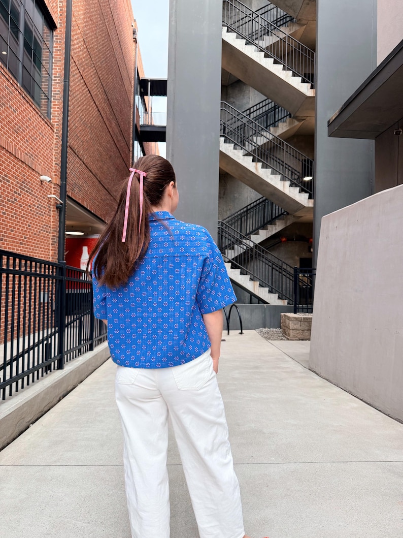 May include: A person wearing a blue patterned short-sleeved shirt and white trousers. The shirt features a floral design. The person's hair is styled with a pink ribbon. The background includes brick buildings and a concrete walkway.