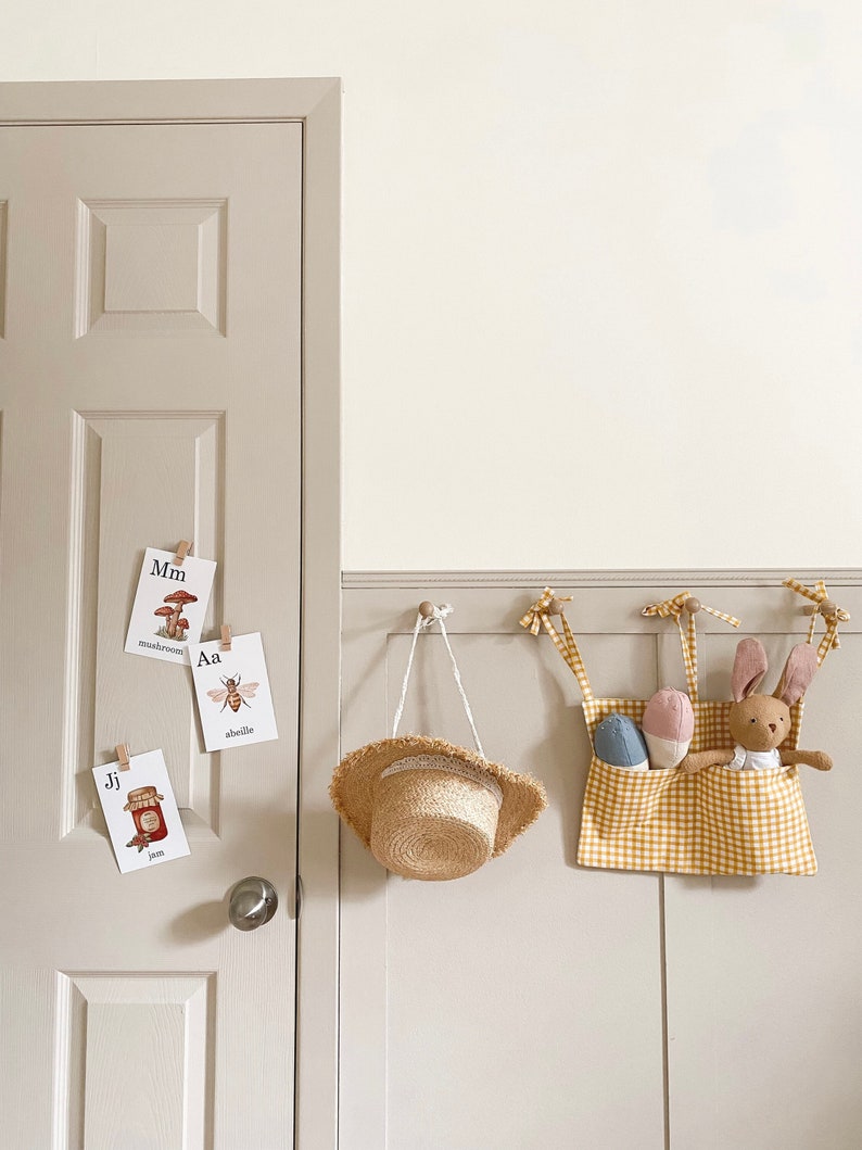 May include: A yellow and white gingham fabric hanging organizer with a stuffed bunny and two stuffed eggs inside. The organizer is hanging on a white wall with a straw hat and alphabet flashcards.