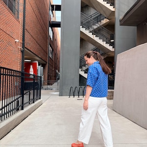 May include: A person wearing a blue patterned short-sleeve shirt, white trousers, and red shoes walks down a concrete walkway. The background features brick buildings, a metal fence, and a staircase.