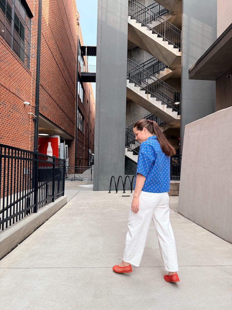 May include: A person wearing a blue patterned short-sleeve shirt, white trousers, and red shoes walks down a concrete walkway. The background features brick buildings, a metal fence, and a staircase.
