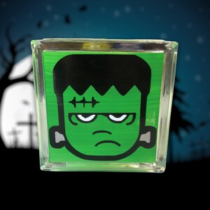May include: A clear glass block featuring a green background with a cartoon illustration of Frankenstein's monster. The monster has black hair, a stitched scar, and gray ear bolts. The background suggests a Halloween theme.
