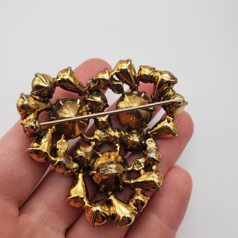 K&ouml;nnte beinhalten: Eine goldfarbene herzf&ouml;rmige Brosche mit einem Cluster aus klaren Strasssteinen. Die Brosche hat eine Stecknadel auf der R&uuml;ckseite und ist auf der R&uuml;ckseite mit "Coro" gekennzeichnet.