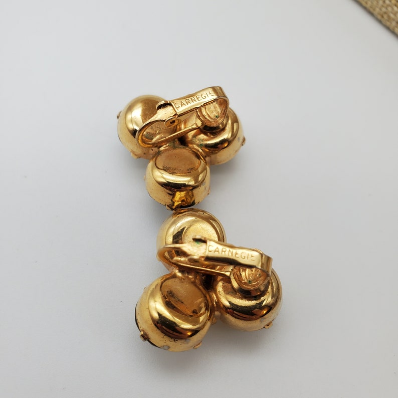 May include: A pair of gold tone clip-on earrings with three round, faceted stones. The earrings are marked "Carnegie" on the back.