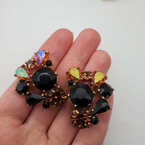 K&ouml;nnte beinhalten: Ein Paar goldfarbene Ohrringe mit schwarzen und schillernden Steinen. Die Ohrringe sind in Form einer Blumenansammlung gestaltet.