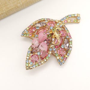 Könnte beinhalten: Eine goldfarbene Brosche in Form eines Blattes mit rosa, blauen, grünen und klaren Strasssteinen. Die Mitte der Brosche zeigt eine Ansammlung von rosa und klaren facettierten Steinen.