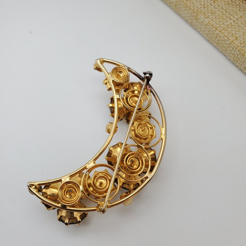 May include: A gold crescent moon brooch with a decorative design of swirling metal and dark blue rhinestones.