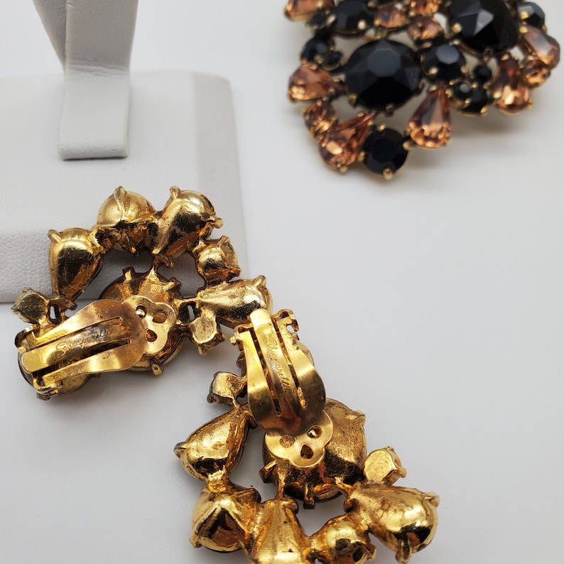 K&ouml;nnte beinhalten: Ein Paar goldfarbene Ohrclips mit einem Cluster aus klaren und schwarzen Strasssteinen. Die Ohrringe sind auf der R&uuml;ckseite mit "Schiaparelli" gekennzeichnet.