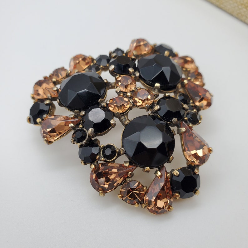 K&ouml;nnte beinhalten: Eine gro&szlig;e, verzierte Brosche mit einem Cluster aus schwarzen und bernsteinfarbenen Strasssteinen. Die Brosche ist aus Metall und hat einen Vintage-Stil.