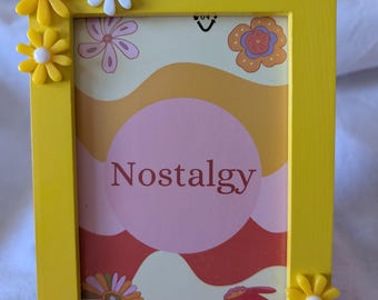 May include: A yellow picture frame with a white and pink floral design inside. The frame is decorated with white and yellow daisy-shaped embellishments.