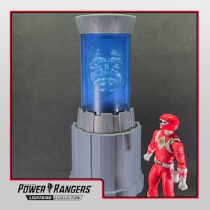 Puede incluir: Un objeto de colección de Power Rangers Lightning Collection con un contenedor teñido de azul con una cara esculpida en su interior. Una figura de acción de Power Ranger roja está cerca. El logotipo de Power Rangers es visible en la parte inferior.