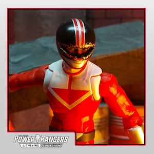 Könnte beinhalten: Rote und weiße Power Rangers Actionfigur mit schwarzem Helm mit roten Streifen. Die Figur hat ein weißes Bruststück mit rotem Pfeildesign und goldenen Akzenten. Das Power Rangers Lightning Collection Logo ist sichtbar.