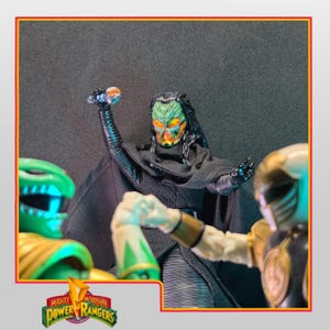 Könnte beinhalten: Actionfiguren von Charakteren aus Mighty Morphin Power Rangers. Eine Schurkenfigur mit einer grün-orangen Maske und einem schwarzen Umhang hält eine leuchtende Kugel. Zwei Power Rangers-Figuren sind im Vordergrund, eine grüne und eine goldene.