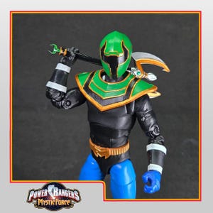 Puede incluir: Figura de acción de un personaje de Power Rangers Mystic Force. La figura es predominantemente negra con detalles verdes y dorados. Presenta un casco verde, hombreras y un arma. La figura posa sosteniendo el arma sobre su hombro.