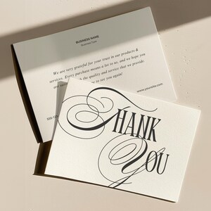 May include: A white thank you card with black calligraphy script that says "Thank You". The card is on a light beige surface.
