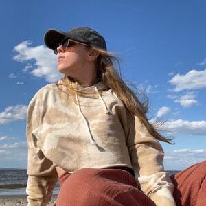 May include: A person wearing a beige tie-dye cropped hoodie with a black baseball cap and sunglasses, sitting on a red blanket on a beach.