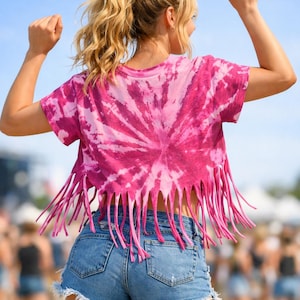 May include: A pink tie-dye crop top with fringe detail and denim shorts. The top features a spiral pattern in shades of pink. The shorts are a light wash denim. The image is taken outdoors on a sunny day.