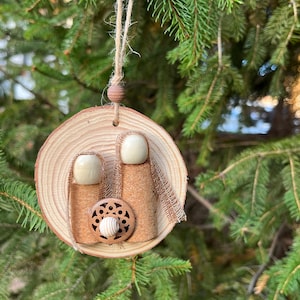 May include: A wooden Christmas ornament featuring a nativity scene. The ornament is round, made of a wood slice, and has three cork figures with wooden heads. A small wooden bead and twine are used for hanging.
