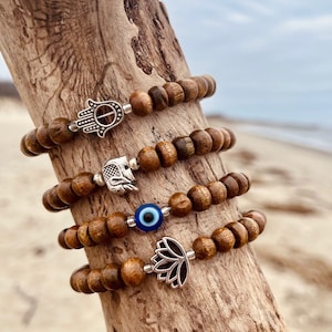 May include: Four wooden bead bracelets with different silver charms. The charms include a hamsa hand, an elephant, an evil eye, and a lotus flower.