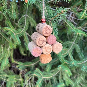 May include: A handmade Christmas tree ornament crafted from repurposed wine corks. The ornament features a cluster of corks in varying shades of brown and red, strung together with twine and a red bead. The corks have the word "MIQUEL" printed on them.