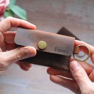 May include: A brown leather key holder with a brass button and a metal key ring. The key holder has the name "Fussell" embossed on the front.