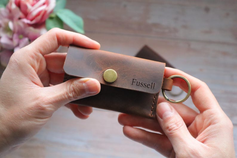 May include: A brown leather key holder with a brass button and a metal key ring. The key holder has the name "Fussell" embossed on the front.