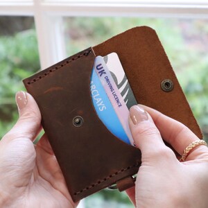 May include: A brown leather wallet with a snap closure and a card slot. The wallet is open and a UK driving licence is visible inside.