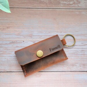 May include: A brown leather key chain with a snap closure and a metal key ring. The keychain is personalized with the name "Fussell".