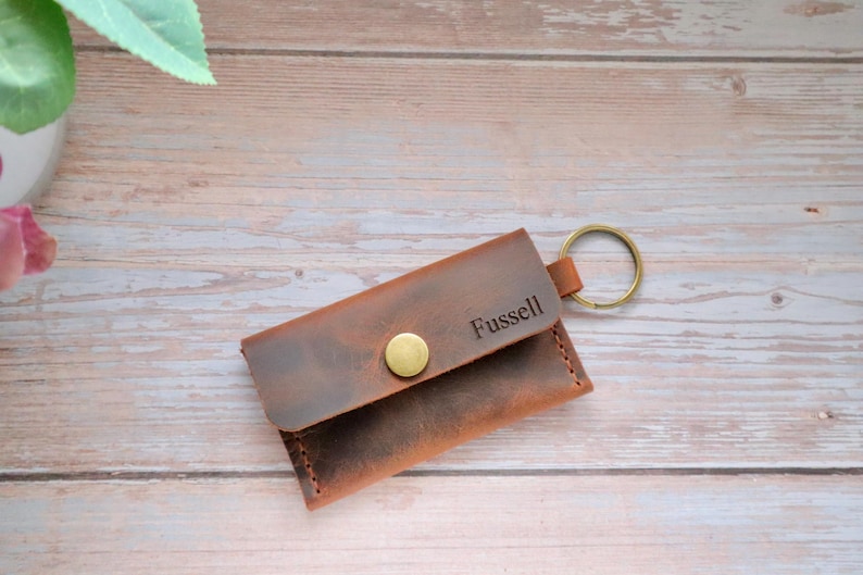 May include: A brown leather key chain with a snap closure and a metal key ring. The keychain is personalized with the name "Fussell".