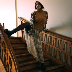 May include: A woman wearing a brown cable-knit turtleneck sweater with leopard print accents on the sleeves, paired with a white lace skirt and black boots. She is positioned on a wooden staircase with a decorative railing.