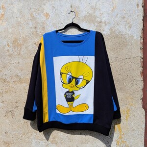 Könnte beinhalten: Ein langärmeliges Sweatshirt mit einem Color-Block-Design in Blau, Schwarz und Gelb. Das mittlere Feld zeigt eine Grafik der gelben Zeichentrickfigur Tweety, die eine schwarze Brille und ein schwarzes Shirt mit dem Text NERDY BIRD trägt.