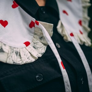Puede incluir: Un cuello blanco adornado con corazones rojos y un ribete de encaje, superpuesto a una camisa negra. El cuello tiene lazos. El ribete de encaje añade un toque delicado al diseño.