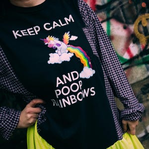 May include: Black long-sleeved shirt with a graphic of a unicorn pooping a rainbow, with the text "KEEP CALM AND POOP RAINBOWS." The shirt has plaid sleeves and a neon yellow skirt.