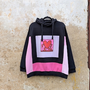 May include: A black hooded sweatshirt featuring a color-blocked design with light pink and hot pink panels. The center of the chest displays a square graphic of a red heart with stick-figure legs and arms, rendered in a bold, cartoon-like style.