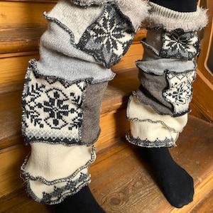 May include: A pair of patchwork leg warmers in shades of gray, cream, and brown. The leg warmers feature a variety of knitted patterns, including snowflake designs. The edges are trimmed with a ruffled detail.
