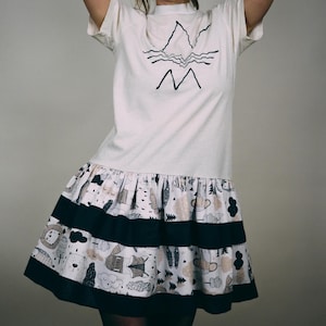 Op de afbeelding: Een witte t-shirtjurk met een zwarte bergafbeelding en een patroonrok. De rok heeft een herhalend patroon van dieren, bomen en wolken in tinten wit, zwart en bruin. Twee zwarte strepen accentueren de rok.