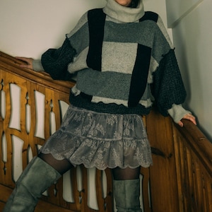 May include: A patchwork sweater in shades of gray and black with a turtleneck collar. The sweater is paired with a gray lace mini skirt and gray knee-high boots. The outfit is worn on a wooden staircase.